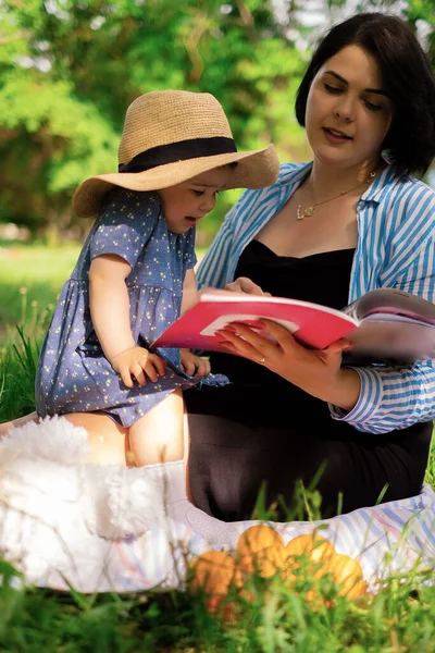 Mom and daughter reading a book on nature Beautiful mother and her cute daughter eat strawberries and tangerines in the park and enjoy a picnic day, family weekend, park vacation