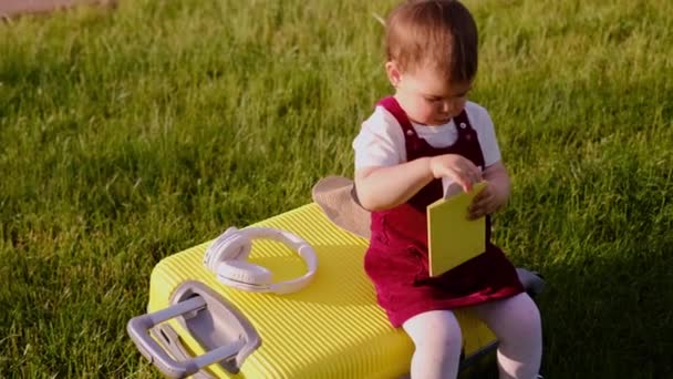 孩子在飞行前坐在行李上 到达后 一个小女孩坐在手提箱上 一个黄色的手提箱躺在绿草上 一个小孩手里拿着车票和护照 — 图库视频影像