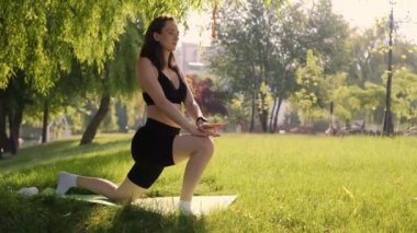 Bir kız, doğa tarafından çevrelenmiş yeşil çimlerin üzerinde lotus pozisyonunda poz verir, yoga ve sağlıklı bir yaşam tarzı için egzersizlerin tadını çıkarır. Doğanın tadını çıkaran genç bir kız parktaki yeşil çimlerde yoga yapıyor.