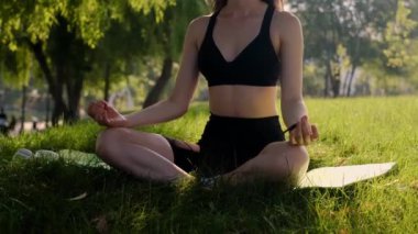 Bir kız, doğa tarafından çevrelenmiş yeşil çimlerin üzerinde lotus pozisyonunda poz verir, yoga ve sağlıklı bir yaşam tarzı için egzersizlerin tadını çıkarır. Doğanın tadını çıkaran genç bir kız parktaki yeşil çimlerde yoga yapıyor.