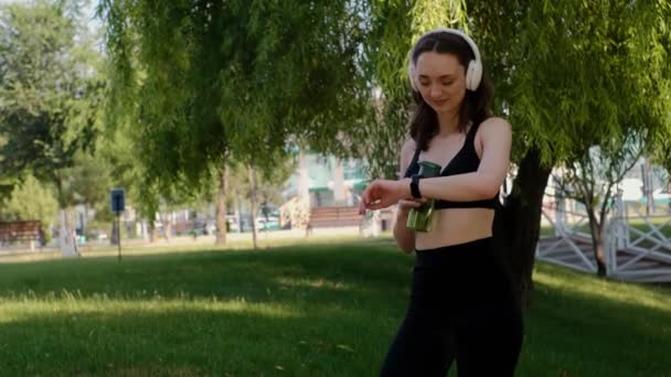 公園でヨガをしている若い幸せな女の子は ヘッドフォンで音楽を聴き 蓮の位置に座って瞑想し バランスよく一人でいることを楽しんで 水を飲んだり 走ったり 公園でジョギングしたりします — ストック動画