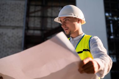 Kasketli mutlu bir mimar inşaat alanında duruyor, elinde planları tutuyor, inşaat alanı yöneticisi koruyucu bir yelek ve miğfer giyiyor ve inşaat alanını düşünüyor..