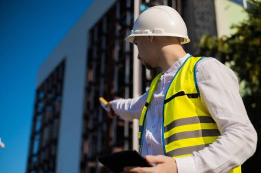 Bir inşaat sahasında çalışırken elini ekranın soluna doğru gösteren bir dizüstü bilgisayarla koruyucu ceketli ve kasklı bir mimarın portresi.