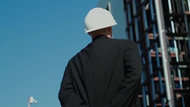 年轻的男性建筑工程师 戴着防护头盔的建筑师工头背着他站在那里 看着多层建筑的建造 城市建筑工地 梦想未来的家园 慢动作 — 图库视频影像
