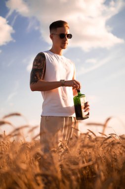 Bir adamın elinde nefis bir alkollü içecek. Bir çiftçi, olgunlaşmış buğday tarlasında taze soğuk bira taşır. Ekolojik içecek, bitkisel karışım, insan içkisi. 