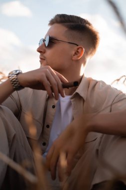 Sahada bej takım elbiseli ve sigara içen güneş gözlüklü zalim bir genç.