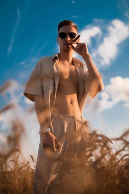 Sahada bej takım elbiseli ve sigara içen güneş gözlüklü zalim bir genç.