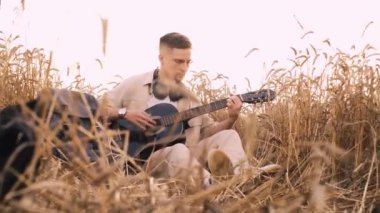 Erkek hipster buğday tarlasında elektro gitarla oynuyor. Uzun saçlı hippi adam arpa otlağında müzik aleti çalıyor. Genç adam doğada dinleniyor. Arka planda güzel bir gün batımı. Yavaş çekim