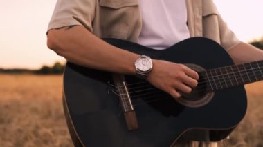Erkek hipster buğday tarlasında elektro gitarla oynuyor. Uzun saçlı hippi adam arpa otlağında müzik aleti çalıyor. Genç adam doğada dinleniyor. Arka planda güzel bir gün batımı. Yavaş çekim
