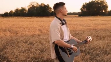 Erkek hipster buğday tarlasında elektro gitarla oynuyor. Uzun saçlı hippi adam arpa otlağında müzik aleti çalıyor. Genç adam doğada dinleniyor. Arka planda güzel bir gün batımı. Yavaş çekim