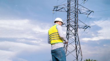 Kasklı ve güvenlik gözlüklü beyaz erkek mühendis, cep telefonu tesislerini kontrol eden telekomünikasyon kulesinin yanında saha çalışması için akıllı telefon kullanıyor..