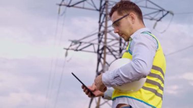 Bir tasarımcı yüksek voltaj kulesini kontrol eder. Bir güç iletim pilonuna hizmet eden kasklı bir mühendis. Güç hattında kask takan bir elektrik mühendisi. Bir tasarımcı yüksek voltaj sensörünü kontrol eder