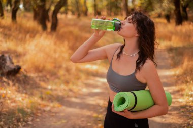 Kadın sonbahar parkında spor yaptıktan sonra şişeden su içiyor, yoga minderi tutuyor, egzersiz yaparken sağlıklı su tüketimi kavramını kullanıyor.