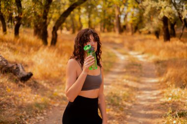 Genç kadın parkta egzersiz yaparken su içer, sağlıklı yaşam tarzı konsepti, doğa ile yalnızlık, sonbahar parkı, ormanda sabah koşusu