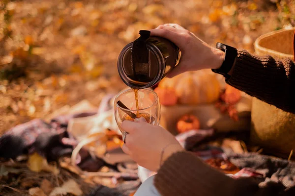 Woman pouring tea from thermos into cup relaxing in autumn forest sitting on trunk. Close up of hot drink and basket with yellow leaves