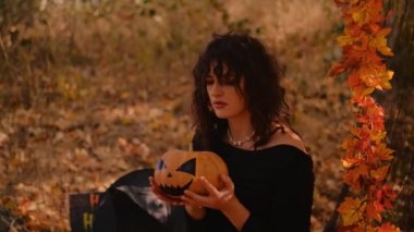 Elinde balkabağı feneri olan cadı bir kız ormanın arka planında poz veriyor. Korkunç suratlı Cadılar Bayramı balkabağı. Cadı kostümü giymiş genç bir kadın. Cadılar Bayramı konsepti Elini sallıyor..