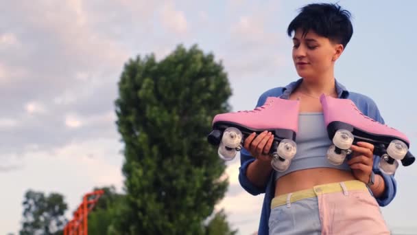 一个留着短发的年轻微笑的女孩手里拿着溜冰鞋 公园里拿着轮滑刀片的女人第一步骤及暑期体育训练概念 — 图库视频影像