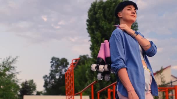 一个留着短发的年轻微笑的女孩手里拿着溜冰鞋 公园里拿着轮滑刀片的女人第一步骤及暑期体育训练概念 — 图库视频影像