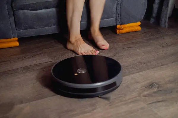 A robot vacuum cleaner cleans the living room. The girl removed her feet to let the robot vacuum cleaner pass. Concept of smart home, technology, housework.