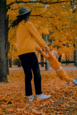Anne oğlunu neşeli bir sonbahar parkında döndürüyor. Aile hafta sonu eğlencesi ve açık havada kaynaşma. Sonbahar mevsiminde aşk ve kahkaha.