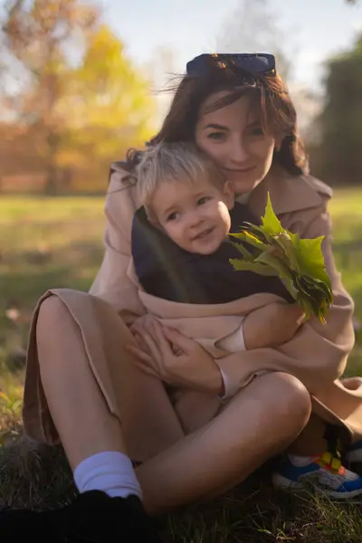 A mother and son embrace and play in the spring park. The boy holds green tree leaves while the mother tickles him, creating precious family moments on Mother\'s Day, enjoying a picnic in nature.