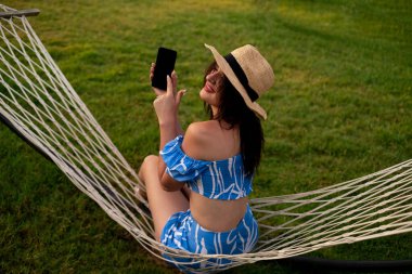 Kız telefonu boş ekranla işaret ediyor, hamağın dışında çalışıyor. Uzaktan çalışma, serbest çalışma, teknoloji, mobil uygulamalar, seyahat, iş aksesuarları, zaman yönetimi ve görev örgütü.