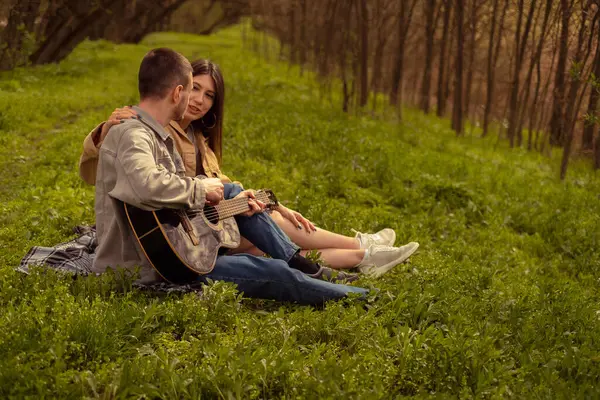 Young couple picnicking in the forest. Man playing guitar, teenage love and friendship, first date, young tourists enjoying outdoor activities.