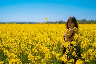 Hasır şapkalı esmer kız, arka planda çiçek açan kolza tohumu tarlasının tadını çıkarıyor, doğanın güzelliğini kucaklıyor..