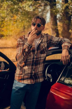 Bir adam arabasının yanında sigara içiyor, egzoz dumanından kaynaklanan hava kirliliğini ve sigara içmenin sosyal ve kültürel etkisini vurguluyor..