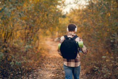 Sırt çantası ve uyku paspası olan bir adam kameradan uzaklaşıyor, ormanın güzelliğinin tadını çıkarıyor, gün ışığını yakalamak için yukarı bakıyor..