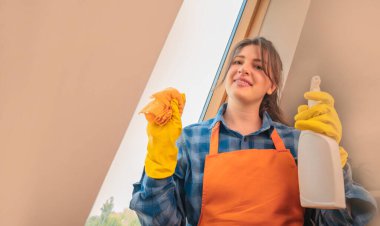 Genç bir kadın dairesinde cam siliyormuş. Elinde bez parçası ve temizlik malzemesi tutuyormuş.