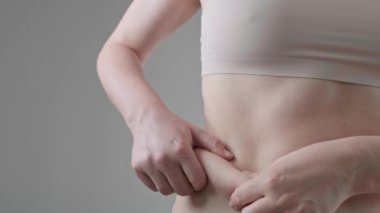 Sırtında gri bir arka planda kalın katlar tutan bej elbiseli genç bir kadının videosu. Şişman, sarkık ve sarkık kaslar. Vücut değişiklikleri