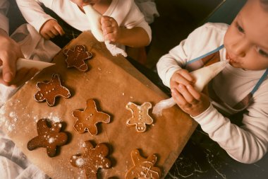 Zencefilli kurabiyelerin olduğu siyah bir masanın üst görüntüsü bir anne ve çocukları tarafından kremayla süslenir..
