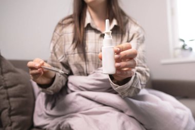 Hasta kadın evde soğuk algınlığını tedavi ediyor, elinde burun spreyi ve doku tutuyor, semptomlara ve evde tedaviye odaklanıyor..