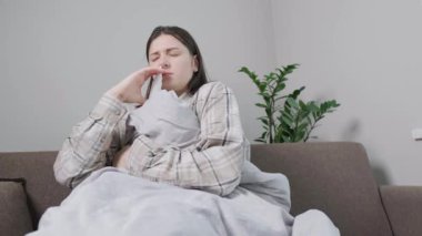 Evdeki kanepede battaniyeye sarılı hasta kadın burun damlası kullanıyor, burun tıkanıklığını deniz suyu çözeltisiyle tedavi ediyor. Yüksek kalite 4k görüntü