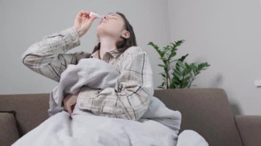 Evdeki kanepede battaniyeye sarılı hasta kadın burun damlası kullanıyor, burun tıkanıklığını deniz suyu çözeltisiyle tedavi ediyor. Yüksek kalite 4k görüntü