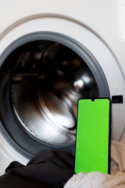 Woman controls the washing machine via mobile phone with Wi-Fi and the Homesmart app, using a green screen interface. clipart