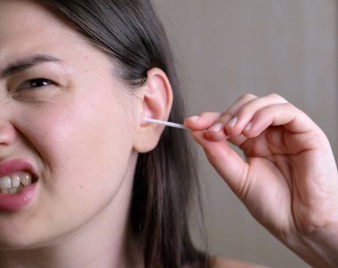 Millennial woman cleaning her ears in the bathroom, feeling pain during ear cleaning and making a grimacing facial expression. clipart