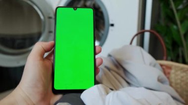 İş önlüklü bir kadın, elinde kroma ekran olan bir telefon, bir çamaşır makinesinin uzaktan kumandası, otomatik işlemler. Yüksek kalite 4k görüntü