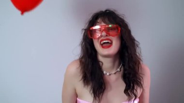 Yukarıdan düşen kalp şeklinde kırmızı balonlarla parlayan kırmızı gözlüklerle dans eden kız Sevgililer Günü partisini kutluyor. Yüksek kalite 4k görüntü