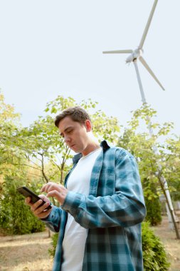 Man taking notes on wind turbine performance, standing in a park with a windmill in the background. clipart