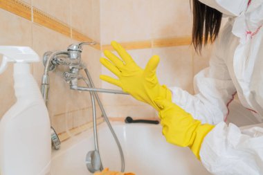 Evde temizlik rutini: koruyucu giysili ve eldivenli bir kadın lavaboya temizleyici sıkar, temizlik ürünleriyle yıkar..