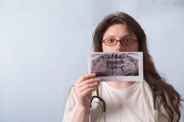 Beyaz önlüklü bir dişçi röntgen görüntüsü tutuyor. Muhtemel diş patolojilerini bulmak için dikkatlice inceliyor..