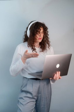 Beyaz kazaklı ve gri pantolonlu öğrenci online derse katılıyor, elinde dizüstü bilgisayarla kablosuz kulaklıkla konferans dinliyor..