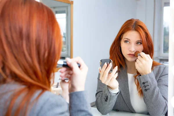 Businesswoman in elegant clothes doing her make up in front of the mirror before going to work. Female empowerment concept