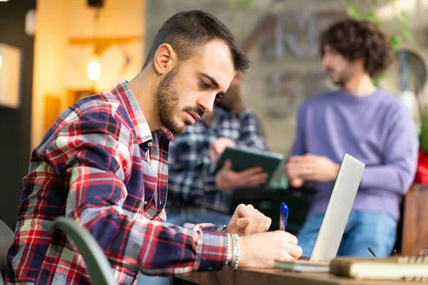 Eş zamanlı çalışma alanında çalışan konsantre bir çocuğa odaklan. Arka planda bulanık çalışan başka adamlar da var. Cafe 'de dizüstü bilgisayarla birlikte çalışma masası.