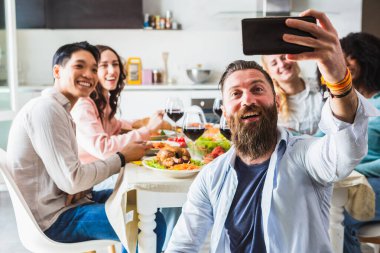 Çok ırklı bir grup gülen genç çocuk öğle yemeği sırasında ev mutfağında grup selfie çekiyor. Yemek yerken gülen ve eğlenen öğrencilerin dairesi.