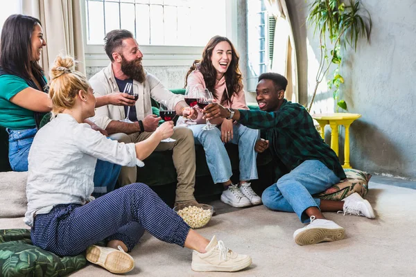Group of friends toasting for home aperitif with red wine sitting on the sofa. Millennial multiethnic people enjoying party staying together. Life style concept with happy guys and girls of different culture