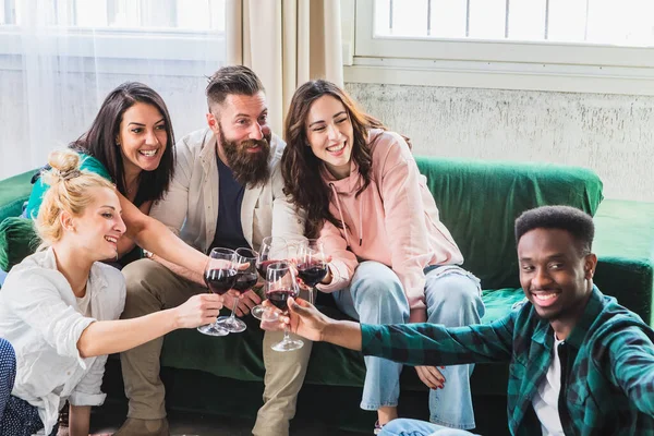 Group of friends toasting for home aperitif with red wine sitting on the sofa. Millennial multiethnic people enjoying party staying together. Life style concept with happy guys and girls of different culture