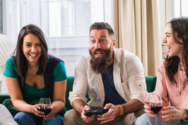 Arkadaşlar gülüyor ve video oyunları oynayarak eğleniyorlar. Konsol kumandalı bir adam ve elinde bir bardak şarapla kanepede oturan iki kız.